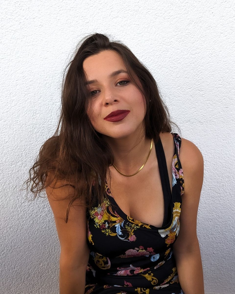 Porträt von Lisa Laner vor einer weißen Wand, mit dunklem, geblümtem Kleid und Goldkette.