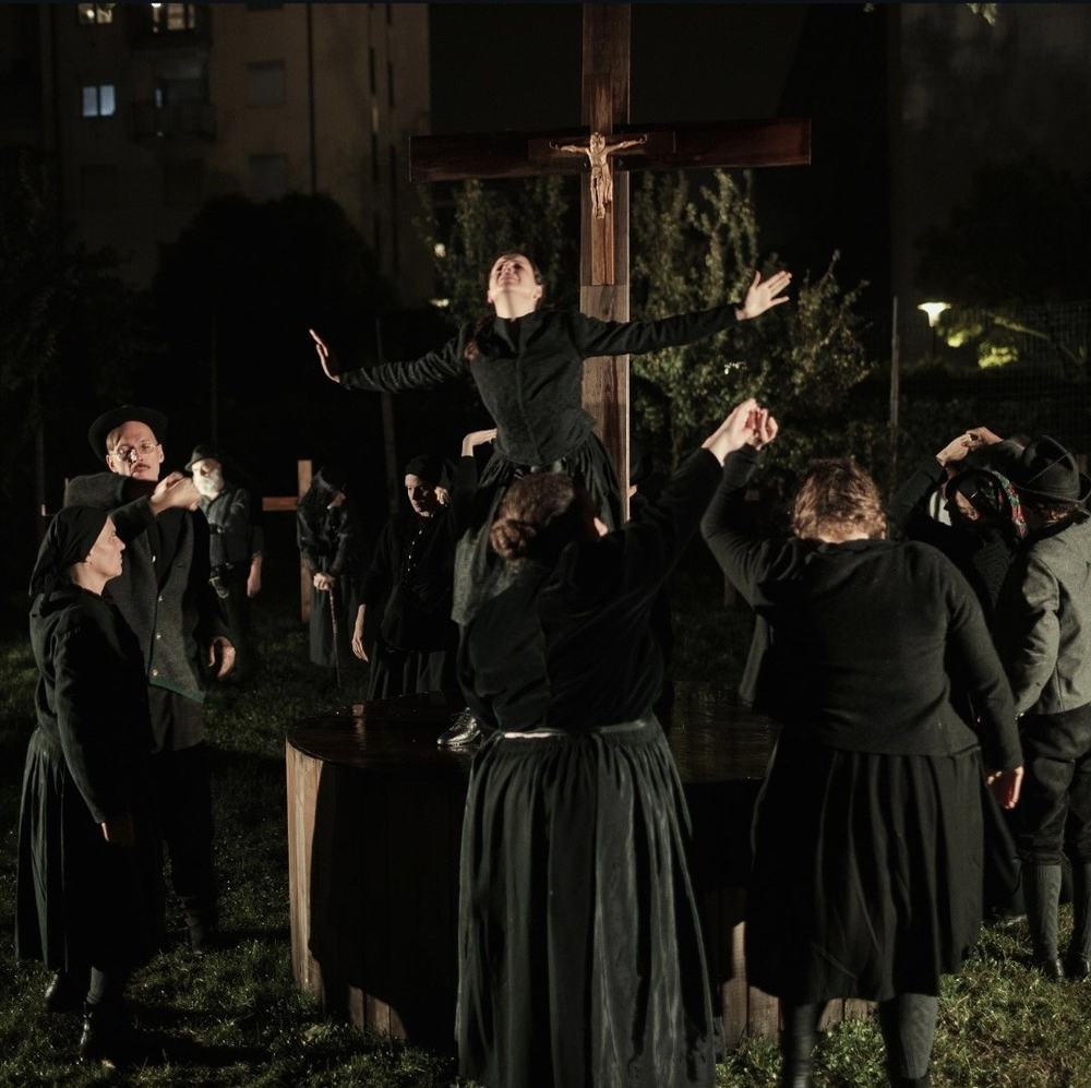Freilicht-Szene bei Nacht: eine Darstellerin steht erhoben vor einem Holzkreuz, eine Gruppe in dunklen Kostümen darunter.