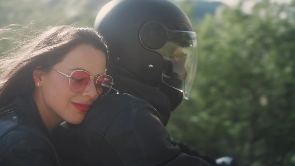 Lisa Laner mit Sonnenbrille auf einem Motorrad, dahinter ein Fahrer mit Helm im Gegenlicht.